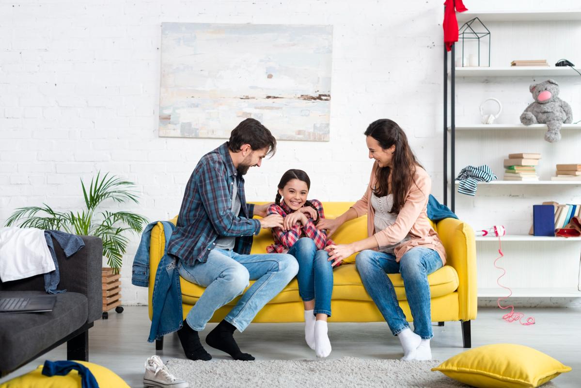 Happy family sitting on a yellow sofa, laughing and playing together in a bright living room with white brick walls and modern decor.