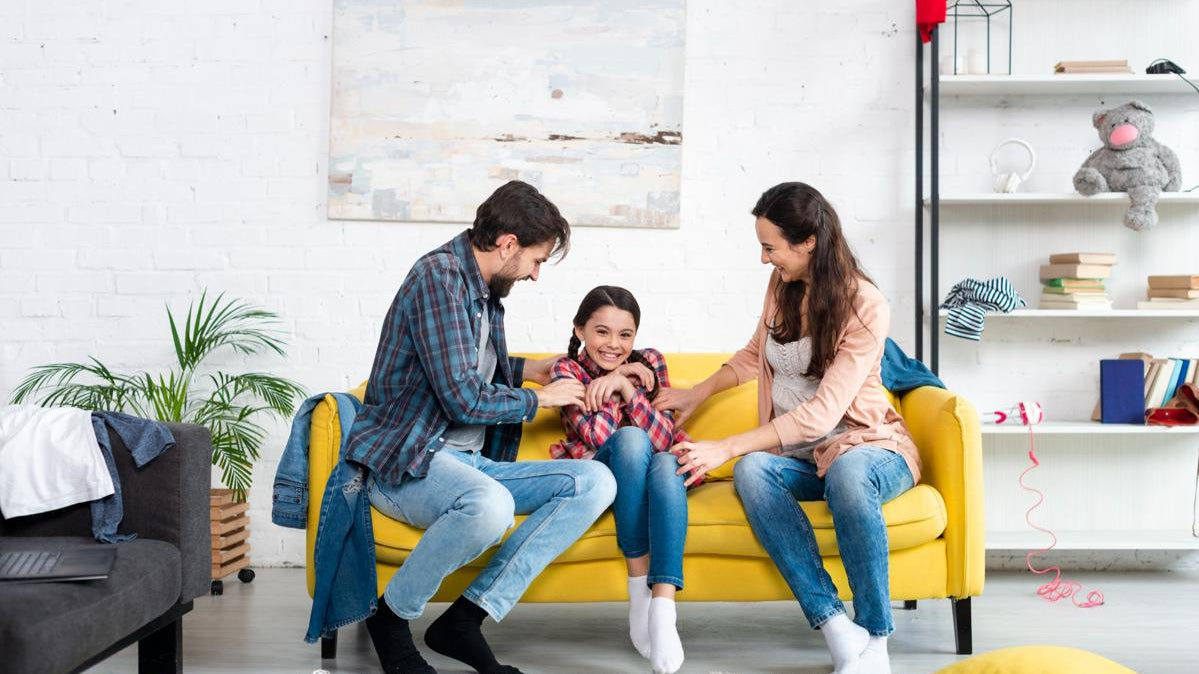 Happy family sitting on a yellow sofa, laughing and playing together in a bright living room with white brick walls and modern decor.
